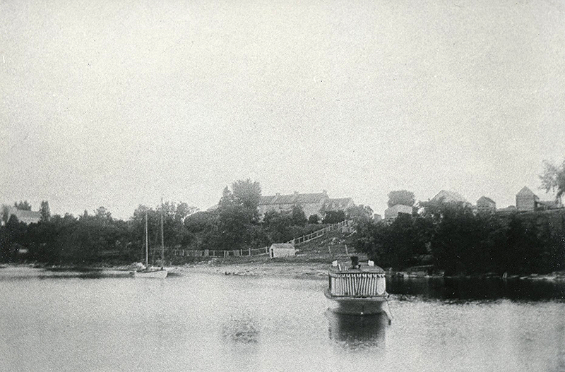 A black-and-white photo of two boats moored in the bay at Pinhey's Point with the Pinhey house and outbuildings and old St. Mary's Church in the background, c. 1900 (Original image in PPF Collections)
